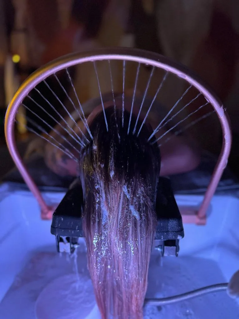 L'arceau d'eau du Head Spa en marche : les filets d'eau tombent sur de longs cheveux mouillés étalés dans le bac, sous une lumière rose et violette.