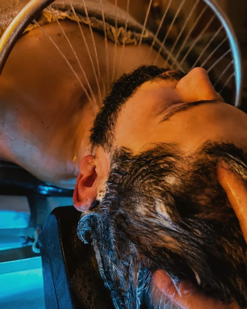 Gros plan sur un homme allongé sous l'arceau d'eau du Head Spa, les mains de la coiffeuse dans ses cheveux mousseux.