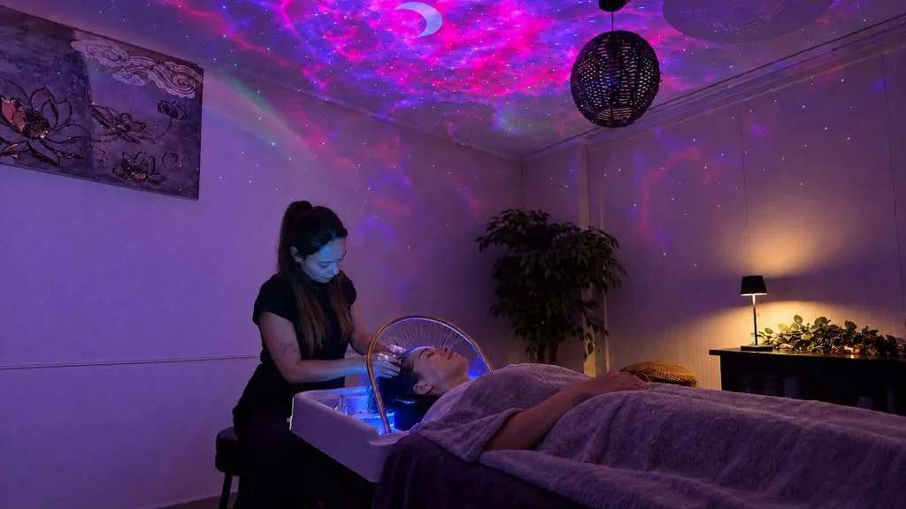 La cabine Head Spa d'Esthé-Tiff à Auvelais : une cliente allongée sous un plaid, la coiffeuse lui masse le cuir chevelu au-dessus du bac, un ciel étoilé projeté au plafond.