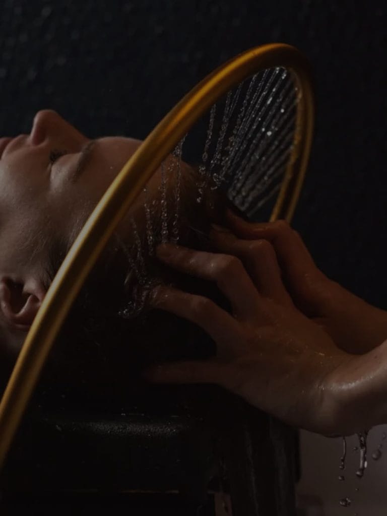 Profil d'une femme allongée sous l'arceau doré du Head Spa, les filets d'eau tombent sur ses cheveux pendant que les mains de la coiffeuse les massent.