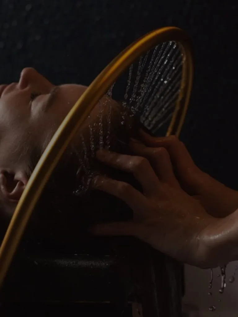 Profil d'une femme allongée sous l'arceau doré du Head Spa, les filets d'eau tombent sur ses cheveux pendant que les mains de la coiffeuse les massent.
